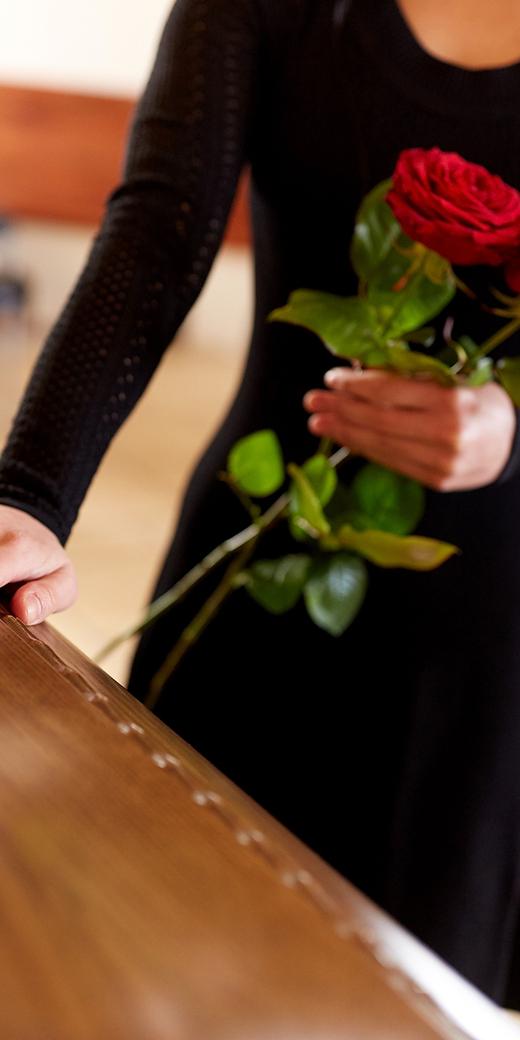 Frau in schwarzem Kleid hält rote Rosen und berührt einen Holzsarg in einem Raum mit Stühlen im Hintergrund.