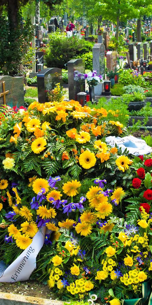 Bunt geschmücktes Grab mit einer Vielzahl von Blumen und einem Holzkreuz auf einem Friedhof umgeben von weiteren Gräbern.