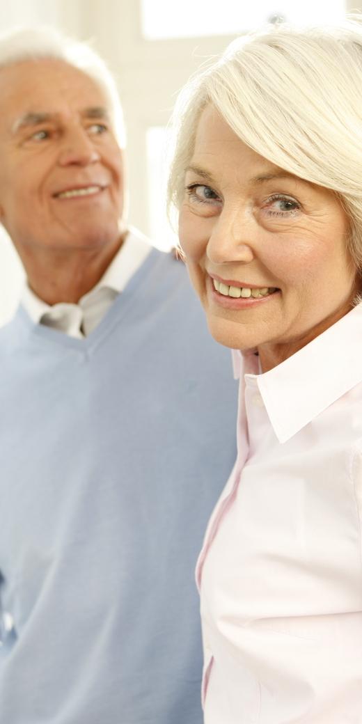Lächelndes älteres Paar steht in einem hellen Raum mit Fenster, die Frau blickt zur Kamera, der Mann schaut sie an.